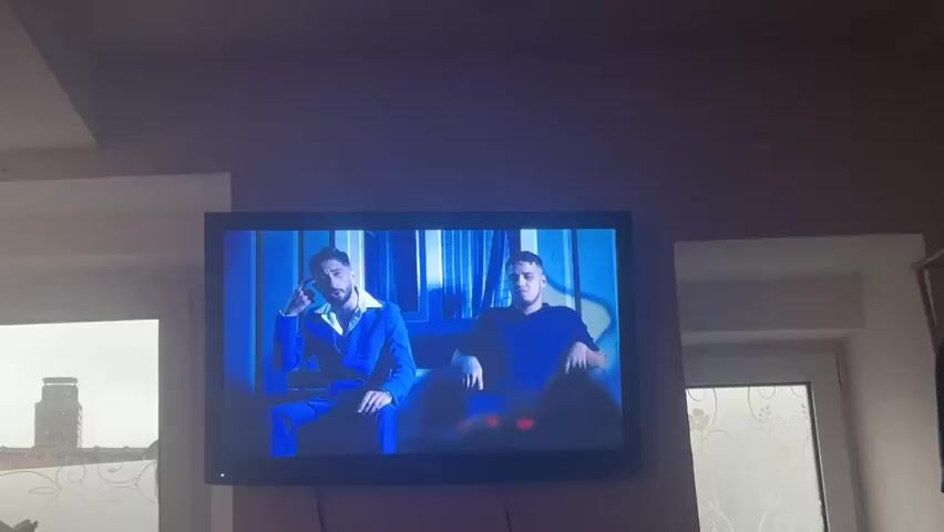 A television is mounted on a wall, displaying two men. One is wearing a blue suit, while the other is in a dark blue shirt. They both appear to be sitting. Two windows flank the television, and the light outside indicates it is daytime. The room's ceiling is white.