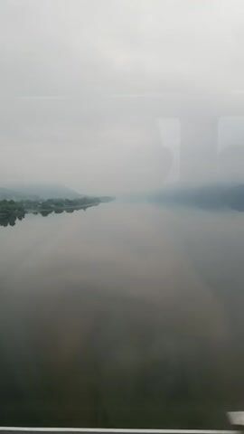 The video shows a wide, calm river with mist or fog obscuring the distant hills. The water reflects the overcast sky, creating a serene and slightly melancholic atmosphere. The view appears to be from a moving vehicle, possibly a train, as indicated by subtle reflections on the glass and the slight motion blur. The left bank of the river shows a strip of greenery with trees and possibly some structures. The overall scene is dominated by muted grays and blues. There are no people or distinct objects visible in the video besides the natural landscape.