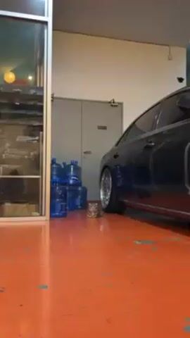 The video shows a small gray tabby cat sitting on a bright orange floor, next to a stack of water jugs and the wheel of a dark blue car. The cat is looking directly at the camera. A rain effect is added over the video. The scene is in a garage-like space with a metal door in the background. The car has shiny silver rims.