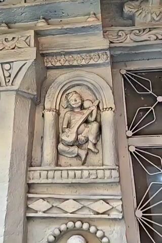 The video features a close-up of intricate stone carvings and architectural details, likely part of a temple in Manikaran, India. The carvings are predominantly light grey or white.

The central focus is a pillar or doorway section with two distinct carvings. The upper carving depicts a figure seated in an alcove, playing a stringed instrument that resembles a sitar or veena. Below it, a larger carving shows a smiling, corpulent figure wearing a crown-like headdress encircled by beads, a beaded necklace, and a 'tilak' mark on its forehead. This figure holds a small pot in one hand, and another pot rests at its feet.

To the left, a decorative archway adorned with vibrant yellow and red marigold garlands is visible. A man, seen from behind, walks through this archway from left to right. He is wearing a plaid vest over a light-colored shirt and pants, and a cap. On the far right, a white-framed window or door with a geometric grid pattern is partially visible. The overall scene showcases traditional Indian temple artistry, accompanied by background devotional music. There is no visible text.
