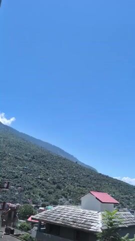 The video pans across a mountainous landscape under a clear blue sky. In the foreground, there are several buildings with pitched roofs, some with traditional slate tiling and others with brightly colored metal roofs, including one prominent red roof. The buildings are clustered together, suggesting a village or town built on the side of a steep, densely forested mountain. The mountain slopes are covered in green trees, with houses and smaller structures visible dotted amongst the foliage. The camera movement is smooth and steady, creating a sweeping view of the settlement nestled within the natural environment. The overall impression is one of a scenic and relatively undeveloped mountainous region.