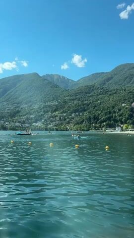 The video shows a wide shot of a lake with mountains in the background. The sky is blue with a few white clouds. The water is a clear turquoise color with gentle ripples. In the distance, there are several people in kayaks or small boats on the water, and yellow buoys mark a section of the lake. The mountains are covered in lush green trees and some buildings are visible scattered on the hillsides. The overall scene is peaceful and suggests a sunny day at a recreational lake.