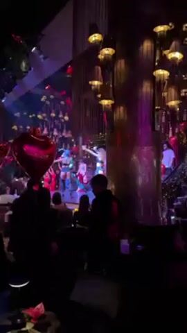 The scene in this video is inside what appears to be a nightclub or event venue. The lighting is low, and the color scheme is dominated by shades of red and purple. There is a stage at the front, where a group of performers is dancing, mostly women, dressed in costumes. Some of the women wear red, while others wear blue. In the foreground, there are patrons seated at tables, watching the show, and a person holding a red heart-shaped balloon. There are also decorative elements like a gold pillar, and elaborate light fixtures hanging from the ceiling.