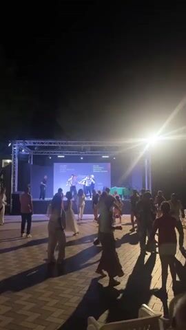 A nighttime outdoor performance is taking place on a stage lit by bright spotlights. A group of performers are on stage dancing and singing. On the screen behind them, text is displayed in white and pink. In front of the stage, a crowd of people are dancing on a paved area. The area is lined with trees.