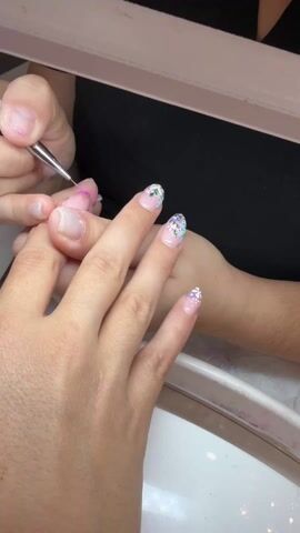 The video shows a person getting a manicure. A manicurist is using a thin, pointed tool to apply small dots of pink polish to the base of the fingernail on the person's left hand. The other nails have already been painted with a design of small flowers and dots in white, purple, and blue. The person's hand rests on a white table near a white basin. The manicurist wears a black shirt. The scene is well-lit.