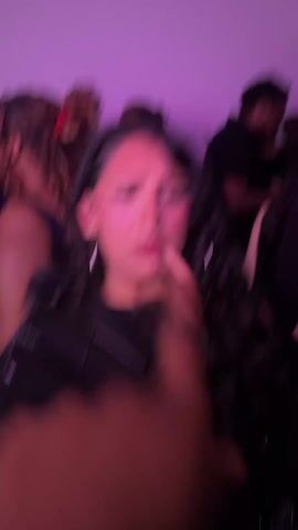 A shaky, handheld video taken indoors, in a room illuminated by pink lighting, captures a young woman looking at the camera with an expressive face, mouth open, possibly speaking or singing. She has long, dark, curly hair, light skin, visible makeup including glitter near her eyes, and multiple piercings, including one on her lip and nose. She is wearing a black jacket and large hoop earrings. In the background, several other individuals are present, mostly dressed in dark clothing, blurred and moving, suggesting a lively social gathering or party. A red plastic cup is briefly visible in the lower left corner of the frame. The environment appears crowded and energetic.