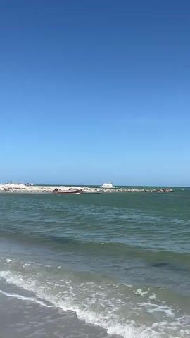 The video shows a view from a beach with calm waves lapping at the shore. In the distance, a breakwater made of rocks is visible. Two boats are docked near the breakwater: a smaller, red boat and a larger, white yacht. The sky is clear and blue.