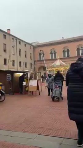 The video shows a plaza on a foggy day. People are walking around a small Christmas market. There is a carousel in the middle of the plaza. There are vendor booths set up along the side of the plaza. There are several buildings surrounding the plaza. The plaza is paved with brick. Some of the buildings are brick and others are yellow. A restaurant with tables and chairs can be seen next to the yellow building.