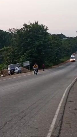 The video shows a gray asphalt road with a white dividing line cutting through the middle. The road is lined with trees on both sides, giving the impression of a rural or semi-rural setting. A white van loaded with items on its roof approaches the camera. In the background, another vehicle can be seen further down the road. A person walks on the left side of the road, heading in the same direction as the van. A sign and a parked vehicle are visible on the side of the road, near a small patch of dirt. The sky appears overcast.