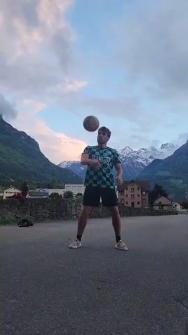A young man stands on an empty asphalt surface, juggling a soccer ball on his head. He wears a green and black checkered t-shirt, black shorts, and tennis shoes. Behind him is a low stone wall, a quaint town with brown houses, and a stunning mountain range with snow-capped peaks under a partly cloudy sky. The man loses control of the ball, which drops to the ground, and he bends over with his hands on his knees, seemingly in frustration. The camera angle is from a medium distance, capturing the man's full body and the scenic backdrop.