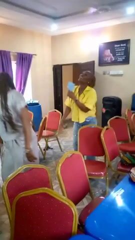 The video shows two women moving chairs in a room. The room has red and gold chairs, and blue tables. One woman is wearing a light-colored dress and has long dreadlocks. The other woman is wearing a yellow shirt and jeans. The room has purple curtains, and a sign that reads "Dear God..." is on the wall. There is a speaker in the background.