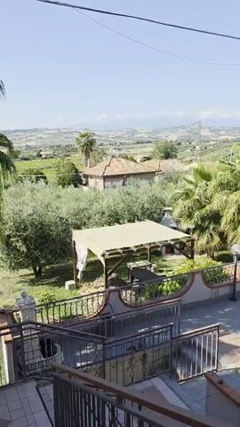 The video captures a scenic outdoor landscape on a bright, sunny day. The camera pans from left to right, revealing a view of rolling hills and distant mountains under a clear blue sky. In the foreground, there is a terraced area with a stone path and a decorative railing, suggesting a patio or balcony. To the left, a wooden gazebo with a light-colored fabric roof stands over a picnic table. Lush greenery, including palm trees, olive trees, and other foliage, fills the middle ground, separating the terraced area from the distant landscape. A few residential buildings with red-tiled roofs are nestled among the trees. The overall impression is one of tranquility and natural beauty, possibly from a countryside or rural setting.