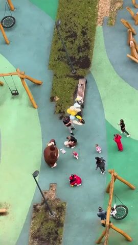 The video shows a playground from an aerial view. There are several people and children in the playground. One person is holding a bunch of balloons. There is a large brown stuffed animal, possibly a bear, in the middle of the playground. Children are playing and interacting with the adults. Some children are sitting on the ground. There are swings and other playground equipment visible. The ground is a patterned surface of green and blue.
