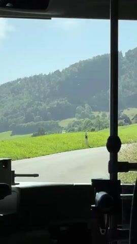 This video captures a scenic view from inside a vehicle, likely a bus, traveling along a road in the Swiss countryside. The scene outside is dominated by rolling green hills covered in lush forests and vibrant meadows. Several scattered farmhouses with red roofs are nestled within the landscape, suggesting a rural setting.

The time of day appears to be late morning or early afternoon, as indicated by the bright sunlight casting shadows and illuminating the landscape. The weather seems pleasant and sunny, with a clear blue sky. The road itself is paved and curves gently through the terrain.

Visible within the vehicle, the interior is partially silhouetted, showing parts of the driver's area. In the rearview mirror, a person wearing glasses and a light-colored shirt can be seen. The overall mood conveyed is one of peaceful travel through a beautiful, natural environment. There is no visible text in the media file.
