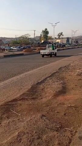 The video presents a wide shot of a street on a sunny day. The road is paved and lined with streetlights. A small truck drives down the street. On the left side of the road, there is a raised, unpaved area covered in reddish-brown dirt. Some vehicles are parked in the background. The camera pans to the right, showing a concrete area near a power pole. In the distance, buildings and some hills can be seen.