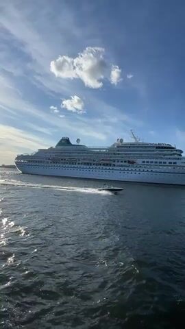 A large white cruise ship is moving through a body of water. A small motorboat is in front of the cruise ship, moving in the same direction. The sky is blue with some white clouds. The water is dark and choppy. The image is taken from a low angle, giving a sense of perspective.