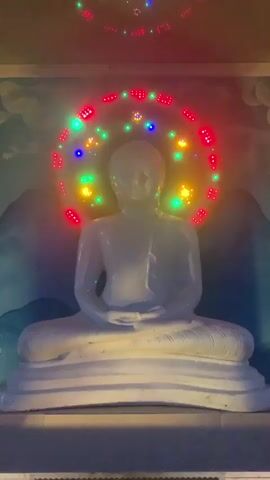 A white statue of Buddha is shown sitting in meditation. A ring of colorful lights, red, green, and yellow, is displayed around the Buddha's head, and the lights twinkle and flash. The statue is set against a backdrop of a blue wall with a painted mountain landscape and white clouds. The lighting in the scene is dim, with the bright lights around the Buddha's head being the primary source of illumination.