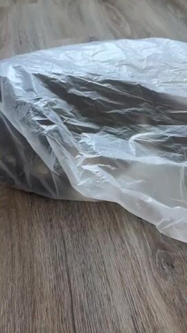 In this close-up, handheld video, a person's finger is visible in the bottom left corner, pointing towards a large, translucent plastic bag. The bag is situated on a wooden floor. Inside the bag, the hind legs and paws of what appears to be a cat are discernible. The cat's fur is black and white, with white paws. The activity captured is a peek into the bag, possibly to observe or interact with the cat. The lighting suggests it is daytime, and no specific weather or emotional cues are evident beyond the playful interaction implied by the presence of the cat and the pointing finger. There is no visible text in the video.