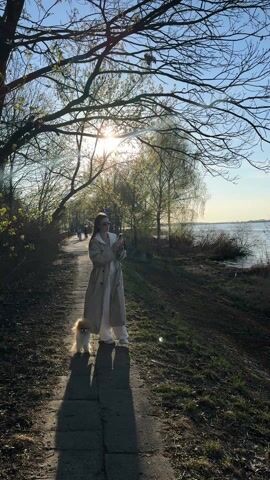 The video features a woman walking a small, fluffy dog, likely a Pomeranian, along a paved path next to a body of water, presumably a river. The setting is a treelined embankment, suggesting a park or natural walkway. The time of day is late afternoon or early evening, indicated by the low sun casting long shadows and creating a bright sun flare through the tree branches in the background. The sky is clear and bright.

The woman is dressed in a light-colored, long trench coat over what appears to be a white top and white wide-leg trousers. She is also wearing sunglasses. As she walks towards the camera, she holds a small device, possibly a phone or a compact camera, in her hands. She momentarily pauses and looks towards the camera before continuing her walk and passing out of frame. The dog walks closely beside her throughout the video. In the background, other people can be seen further down the path. The vegetation along the path includes trees with some new spring growth and others still mostly bare, alongside grassy areas. The river is calm, reflecting the clear sky. No visible text is present in the video. The location is Białobrzegi, Poland.