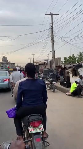 The video, taken from the back of a moving motorcycle, shows a street scene in what appears to be a bustling neighborhood. The motorcycle has a Nigerian license plate. Various individuals can be seen, including pedestrians carrying items, and people sitting or standing near what appear to be shops or market stalls. Objects visible include parked motorcycles, cars, water tanks, furniture, and general street clutter. Overhead, a network of electrical wires dominates the sky. The weather appears overcast.