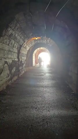 The video shows the interior of a dark, arched tunnel with rough, textured walls. The ground appears to be asphalt.  In the distance, at the end of the tunnel, there is a bright light, suggesting an exit. Two figures, silhouetted against the light, are walking away from the camera towards the exit.  There are some metal pipes and wires visible on the ceiling of the tunnel, and what appears to be a single light fixture hanging from the ceiling.
