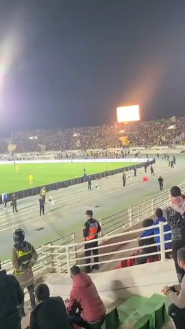 The video shows a sports stadium at night, filled with spectators. A soccer field is visible with players and officials on it, and a large screen displays an advertisement.  There is a track surrounding the field, and security personnel are stationed between the track and the stands. The camera is positioned within the stands, capturing a view of the crowd and the game. Some people in the stands are standing and looking towards the field.