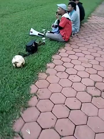 A group of young men are gathered by the edge of a soccer field, observing a game. Four individuals are seated on a hexagonal paved path next to the green grass, with one man in a red and white shirt and a grey cap speaking into a phone. A megaphone, a black bag, a soccer ball, and several cups are placed on the grass near them. In the background, a soccer game is in progress on the large grassy field, with players visible near a goal. The scene is outdoors, surrounded by trees and some buildings in the distance, suggesting a park or recreational area during the daytime, likely late afternoon.