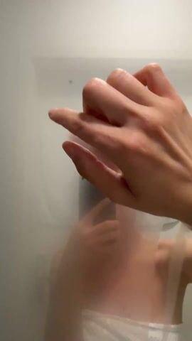 The video shows a woman taking a selfie in a steamy shower. She is holding a phone and looking at it, partially obscured by the condensation on the shower door. She is wearing a towel, covering her upper body. A hand is seen drawing on the steamy glass above her.