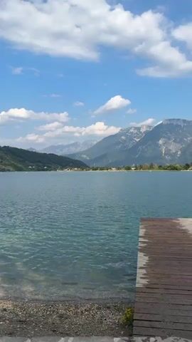 A vertical video displays a serene lake scene under a bright blue sky with scattered white clouds. The water is a clear, tranquil blue-green, reflecting the sky and surrounding mountains. The foreground shows a shoreline with pebbles and sand, transitioning to a paved walkway. Several paddleboards are visible resting on the shore near the water's edge. In the distance, rolling green hills and majestic mountains form a picturesque backdrop. A few small boats and people swimming or wading in the lake can be observed. The overall atmosphere is one of peaceful recreation and natural beauty on a sunny day.