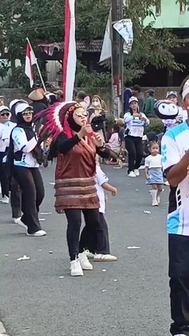 A group of people are participating in an outdoor parade. The central figure is a woman wearing a headdress resembling Native American war bonnets, a brown vest with fringe detailing, black leggings, and white sneakers. She is also wearing sunglasses and makeup. To her left, several women in white tops and black pants and headscarves are marching in unison. To her right, a small child in a white shirt and black pants is also moving. In the background, there are spectators sitting on the side of the road, and red and white flags are visible. The parade appears to be taking place on a paved street.
