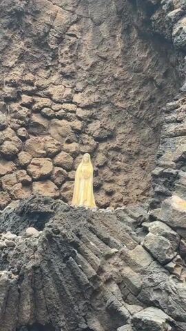 The video shows a cream-colored statue of the Virgin Mary placed atop large, jagged, gray rocks. The statue stands out against the backdrop of the rocks, which display a variety of textures and cracks. A small green and yellow cloth is nestled on the rocks near the statue. The video pans upwards along the rocky cliff face, showcasing the rough, uneven surface of the stone.