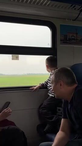 Inside a train, a young boy in a striped shirt sits on a backpack, looking out the window at a green, flat landscape. Next to him, an older man watches the same view. Another man is partially visible, looking at a phone. There's a framed picture of a building mounted on the wall above. The train window is cloudy.