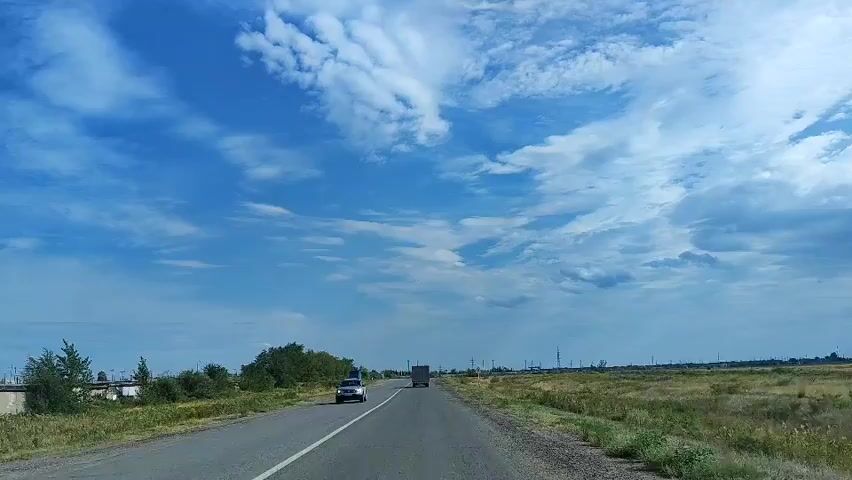The video shows a highway scene. The camera is mounted on a vehicle, suggesting a point-of-view shot from a driver or passenger. The road is paved and appears to be a two-lane highway with a white dividing line. On the left side of the road, there are some scattered buildings and trees. On the right side, there is a grassy field with some dry vegetation. The sky is blue with scattered white clouds. Two vehicles are visible on the road: a white car in the left lane and a truck in the right lane, both moving away from the camera. Traffic signs are visible on the right side of the road. The overall environment suggests a rural or semi-rural setting.