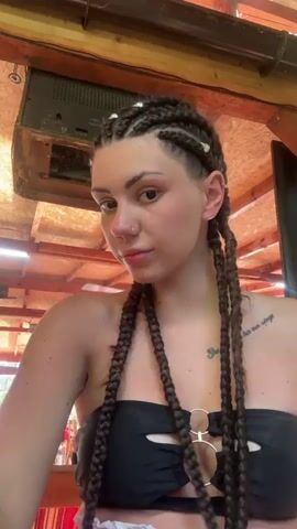 A young woman with braided hair and a black bikini top poses for a selfie. She is indoors, with exposed wooden beams and a ceiling-mounted speaker visible in the background. She moves her hands, touches her braids, and puckers her lips.
