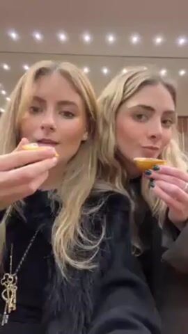 Two young blonde women are eating custard tarts. The camera is held up close as they each take a bite of the tart. The woman on the left is wearing a black top with a feather boa and a long key pendant. The woman on the right is wearing a brown jacket and has dark green nail polish. They appear to be inside a building with a decorative ceiling of small lights.