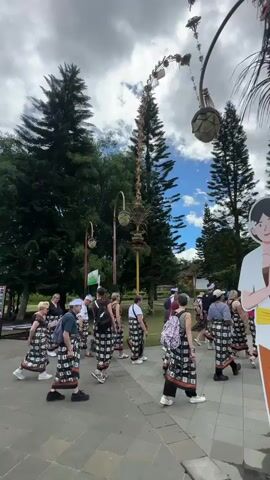A group of people, dressed in a mix of casual and traditional clothing, are walking along a paved pathway. Many are wearing sarongs, a traditional Indonesian garment, and some are wearing white headbands. They are walking away from the camera, heading towards a traditional Balinese structure. The environment includes tall trees, manicured grass, and ornate archways with hanging decorations. The sky is cloudy, with occasional patches of blue visible. A large decorative archway is visible in the foreground. The people seem to be part of a tour group, exploring the area. The camera follows behind them as they walk.
