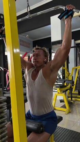 A man with blonde hair and a mustache is performing a lat pulldown exercise in a gym. He is wearing a white tank top and blue shorts. He is holding a bar attached to a cable machine with both hands, pulling it down towards his chest. He is seated on a padded bench with his legs secured under a padded bar. Sweat is visible on his tank top and his face. There are other gym equipment visible in the background, including weight machines. A bottle of water is on a surface to the left of the man. The lighting in the gym appears to be artificial.