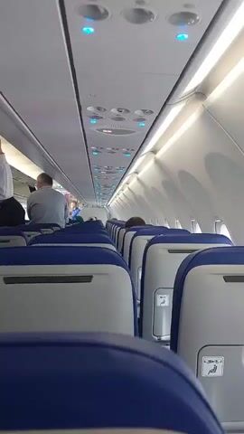 The video shows the interior of an airplane cabin. Passengers are settling into their seats. The seats are blue and gray, and the cabin is brightly lit. A man puts on a white hoodie, and a woman behind him puts on a gray jacket. The overhead compartments are closed. An announcement is being made over the plane's speakers.
