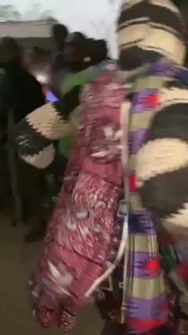 A group of people are gathered in what appears to be an outdoor event. Many are dressed casually, while one individual stands out in a colorful, patterned outfit, including a distinctive striped and patterned hat. This person is moving energetically, possibly dancing. Some people are taking photos or videos with their phones. The background suggests a village setting, with trees and buildings visible. The weather seems overcast, contributing to the slightly muted lighting of the scene.