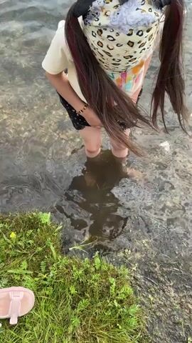 A young girl with long, dark hair in pigtails stands in shallow, clear water at the edge of a body of water. She is wearing a light-colored t-shirt with colorful graphics, dark shorts, and a leopard-print bandana or headband. Her feet are submerged in the water, and she is looking down at them with a slight smile. The water is gently rippling, and the bottom is visible with small stones and pebbles. To the left, there is a patch of green grass with some small yellow flowers. A pink sandal is visible on the grass in the foreground. The lighting suggests it is daytime, likely sunny. The overall scene appears to be a peaceful and enjoyable moment of a child interacting with nature.