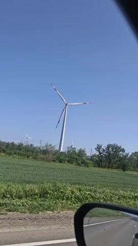 The video, taken from inside a moving vehicle, shows a landscape featuring a wind turbine in a field against a bright blue sky.  The camera angle is initially focused on the turbine, then shifts to include the car's side mirror and the road. The foreground consists of green fields dotted with trees. The driver's perspective includes the steering wheel and dashboard, which displays some metrics related to the drive. The car is moving along a paved road.