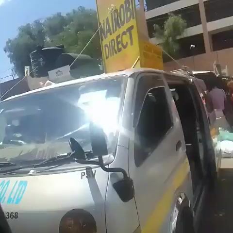 The video shows a view from inside a vehicle in what appears to be a busy urban area, likely in Kenya based on the signage. The first part of the video shows several passenger vans, called "matatus," which are colorfully decorated and bear the names of different travel companies. The video then moves to show the streetscape, which includes pedestrians, motorcycle taxis ("boda bodas"), storefronts with prominent branding (e.g., Safaricom, Tecno, Infinix), and general street activity. The buildings are a mix of architectural styles, and the weather is sunny. The camera person is likely a passenger in the vehicle, as the view is from the inside looking out through the window. The vehicle seems to be moving slowly through the street.