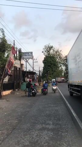 This is a vertical, eye-level, outdoor, daytime video showing a street scene. The pavement is gray and cracked. On the left side of the street, there are buildings and a sign that reads "SMPN 1 GEMPOL". A red and white motorcycle is parked. A person wearing a red helmet and a blue backpack is standing next to it. Another person on a motorcycle wears a black helmet and a gray backpack. To the right of the street, a large white semi-truck is driving away from the camera. Three motorcycles are in motion on the street, with riders wearing helmets. The sky is a pale blue with scattered clouds. Trees and utility poles are visible in the background.
