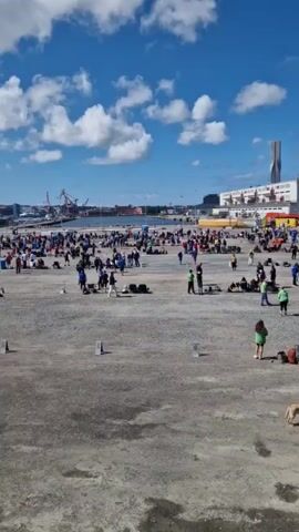 The video shows a large outdoor gathering of people on a gravel surface with a concrete edge in the foreground. In the background, there are several buildings, including a modern skyscraper-like structure and a building with a distinctive patterned facade. Cranes are visible in the distance, suggesting a port or industrial area. The sky is blue with scattered white clouds. People are scattered across the gravel area, some standing, some sitting, and some with luggage or bags. There are also some dogs present. The overall impression is of a public event or gathering, possibly a race start or finish, or a transportation hub. The lighting suggests it is daytime with clear weather.