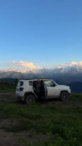 A white Jeep is parked on a grassy hillside with its driver's side door open. A person, seen from behind and with their back to the camera, is reaching into the Jeep. In the background, a vast mountain range with snow-capped peaks stretches across the horizon under a clear blue sky with scattered clouds. The foreground is dominated by green grass and some out-of-focus plants.