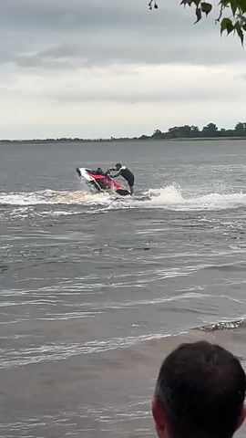 The video shows a person on a jet ski in a body of water. The jet ski is black and red. The person is wearing a light-colored shirt and dark shorts. The person is standing on the jet ski and appears to be performing a maneuver, as the jet ski is tilted to one side and creating a wake. The water is choppy, and the sky is overcast. In the foreground, the back of someone's head is visible, indicating that the video is being filmed from the shore or from another boat. The background shows a shoreline with trees and a flat horizon. The video is approximately 5 seconds long.