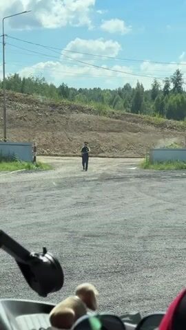 The video shows a person wearing a dark t-shirt, dark pants, and a dark hat walking on a gravel surface. The person is holding a white object in their right hand and appears to be looking at it. The background consists of a dirt embankment covered in sparse vegetation and trees, with a blue sky and white clouds overhead. In the foreground, a black object, possibly a rearview mirror, and other indistinct objects are visible. The person is moving from right to left across the frame.