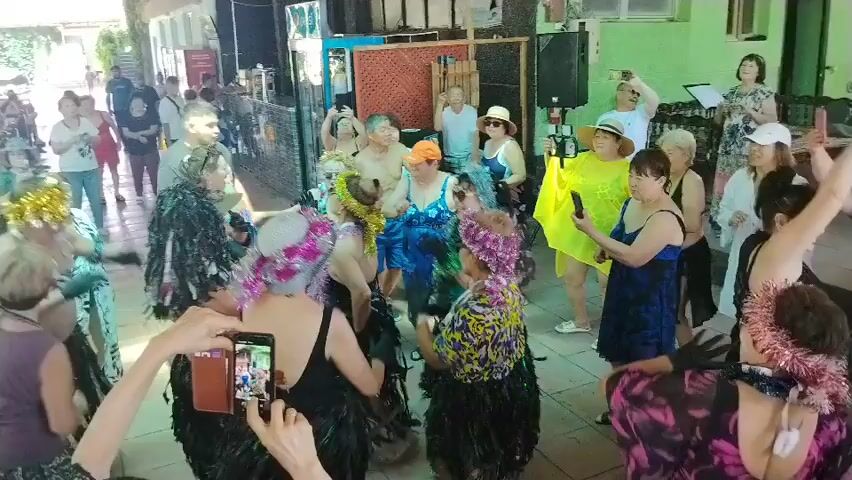 A group of people are gathered in an outdoor, shaded area. Some are dancing in the foreground, wearing floral leis and grass skirts, while others watch or record the event with their phones. In the background, more people are observing, and there is a stage with a sound system. The overall atmosphere appears festive.