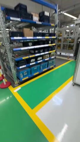 The video opens with a blurry shot of a modern industrial environment. The floor is a light gray with distinct green and yellow safety markings. To the left, shelves filled with various items are visible, also appearing blurred due to motion. The camera then pans to the right, focusing on a large digital screen mounted on a dark blue and lime green structure.

The screen displays an animated graphic. On the left, a cartoon woman wearing a blue cap and green overalls stands next to a thermometer-like gauge labeled "ENERGY." The gauge shows different levels: "low," "medium," and "high," with a red indicator pointing towards the "medium" range. Below the graphic, text in Dutch reads: "Ga na of machines en apparatuur met minder stroom kunnen worden gebruikt of of te kunnen worden uitgeschakeld wanneer ze niet worden gebruikt." This translates to: "Check if machines and equipment can be used with less power or can be switched off when not in use." The screen also has a logo on the bottom right, and a section on the top right of the structure shows the words "WE ARE." The background of the industrial space behind the screen is again visible, with more shelving and lighting. The video appears to be a brief walkthrough or presentation within a facility, likely related to energy efficiency or operational procedures.