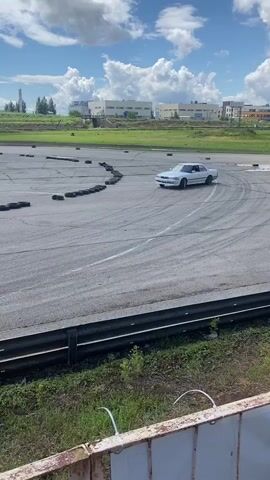 A white sedan is drifting on a paved track. The car is kicking up smoke from its tires as it slides around a corner. There are several black tire obstacles placed around the track. In the background, there are buildings and a cloudy blue sky. The video is shot from a slightly elevated position, looking down at the track.